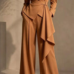 A model wears a camel brown wrap-style jumpsuit with wide legs, a matching waist tie belt, and dramatic layered ruffle drapes on the front. The image is cropped from the chest down, set against a warm, minimalist beige studio background with soft shadows.