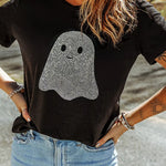 A woman wearing a black short-sleeve t-shirt with a rhinestone-embellished ghost graphic on the front, paired with light blue jeans, standing outdoors.