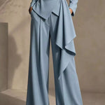 A model wearing a light blue tailored jumpsuit with a belted waist, long sleeves, and wide-leg draped trousers, set against a neutral beige studio background.