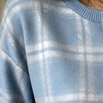 A close-up of a person wearing a light blue crewneck sweater with a textured, metallic-looking plaid pattern in white and light blue. The person is also wearing multiple delicate gold chain necklaces.
