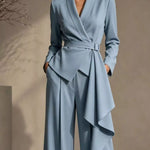 A woman models a light blue tailored jumpsuit with a wrap-style waist, belted tie, long sleeves, and draped asymmetrical skirt panels, photographed against a warm beige wall.