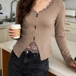 A woman in a kitchen wearing a beige long-sleeve button-up cardigan with a lace V-neck and hem, paired with dark gray jeans and holding a coffee cup.