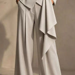 A close-up of a model wearing high-waisted, wide-leg trousers in a light warm gray or greige color. The pants feature a dramatic asymmetrical draped wrap detail on the right side and a matching belt at the waist, set against a neutral textured beige background.
