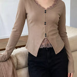 A woman wearing a beige long-sleeve cardigan with a scoop neckline, lace trim, and front buttons, paired with black pants, standing indoors next to a sofa.