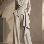 A woman models a light gray wrap-style blazer with matching wide-leg trousers in a minimalist beige-toned studio setting.