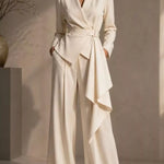 A woman models a cream-colored wrap blazer with matching wide-leg trousers and a self-tie belt, styled in a minimalist beige studio setting.