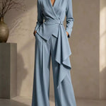 A woman models a light blue tailored jumpsuit with a wrap-style bodice, belted waist, draped asymmetrical skirt detail, and wide legs in a softly lit indoor setting.