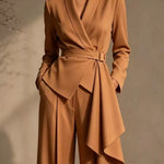 A woman wearing a camel-brown tailored jumpsuit with a wrap-style blazer bodice, belted waist, draped asymmetrical skirt, and wide legs, photographed against a warm neutral wall.