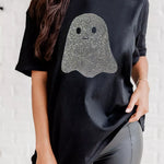 A woman wearing a black short-sleeve t-shirt with a glittery ghost graphic on the front, paired with gray leggings, standing against a white brick wall.