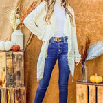 Woman in a stylish outfit with a white cardigan, blue jeans, and black boots standing in front of a wooden crate with decorative items.