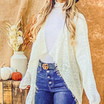 Woman wearing a white cardigan, blue jeans, and a navy hat against a wooden background with pumpkins and a vase.