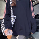 A person wearing an oversized black sweatshirt with pink stripes down the sleeves, paired with gray sweatpants and a black-and-white checkered shoulder bag, standing near a car door.