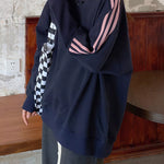 A person wearing an oversized navy sweatshirt with pink stripes on the sleeve, a black-and-white checkered side panel, and dark gray pants, standing indoors against a rustic wall and wooden bench.