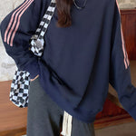 A person wearing an oversized navy sweatshirt with pink stripes on the sleeves, paired with dark gray drawstring sweatpants and a black-and-white checkered shoulder bag, standing indoors against a rustic wall and wooden bench.