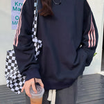 A person wearing an oversized navy sweatshirt with pink stripes down the sleeves, paired with gray sweatpants and carrying a black-and-white checkered tote bag while holding an iced drink.